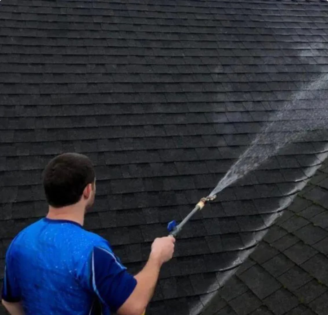Worker in blue shirt pressure washing dark roof shingles with spray wand