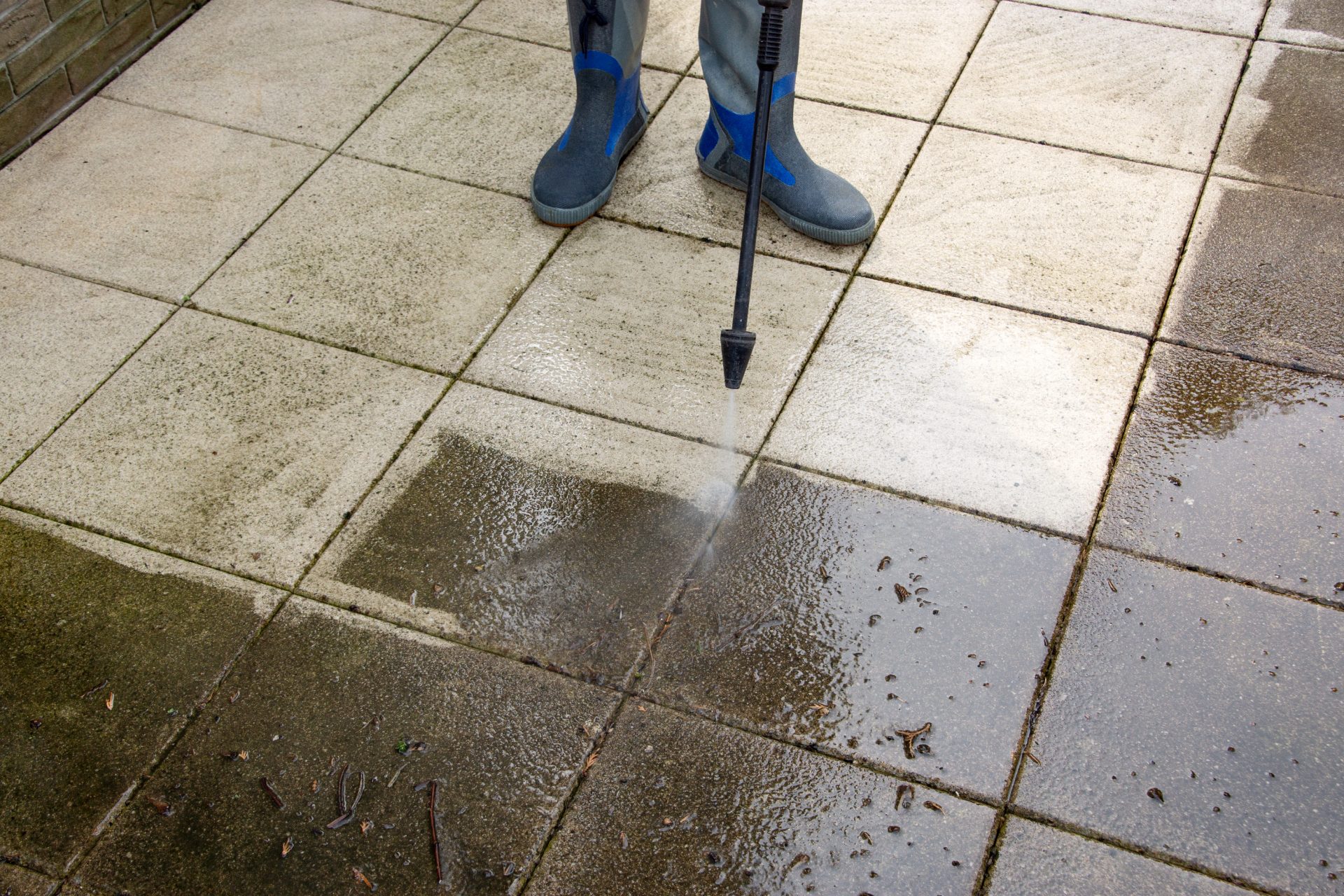 Person in gray and blue boots pressure washing dirty patio tiles with a lance