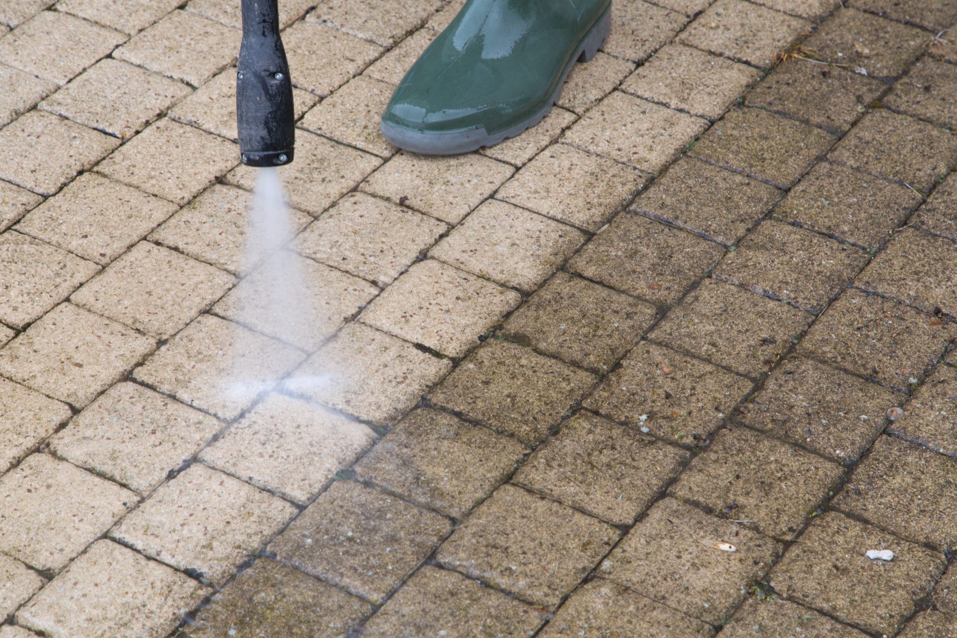 Person pressure washing brick pavers with green boots visible